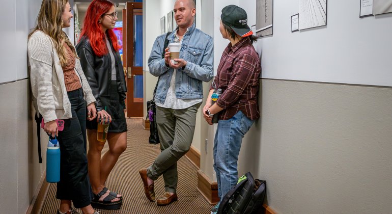 Three students have an informal discussion with a professor in the hallway.