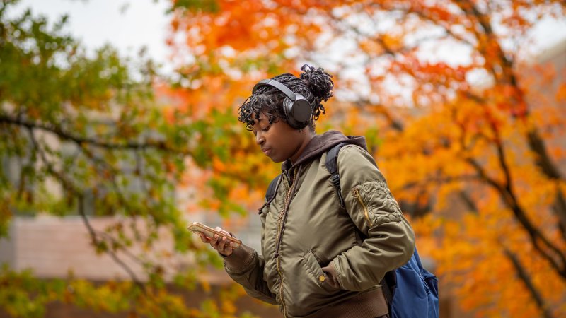 A student walking while looking at her phone with autumn trees behind them