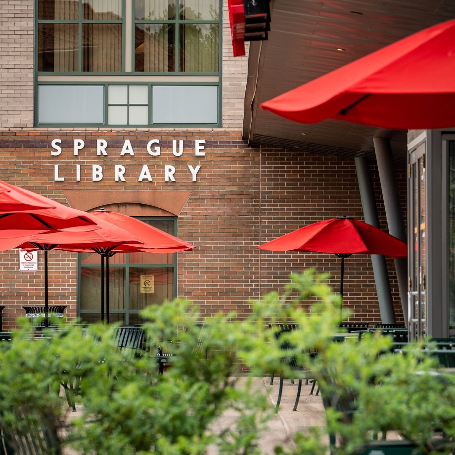 exterior view of Sprague Library showing red umbrellas over round picnic tables