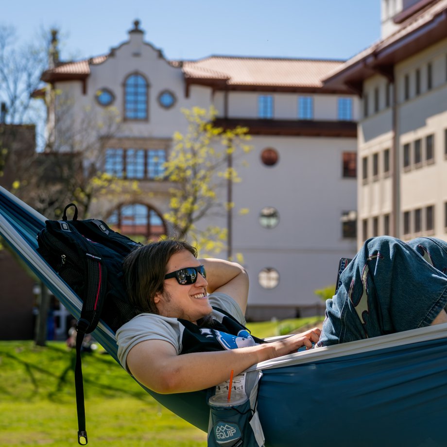 student smiling at computer on hammock in front of university hall