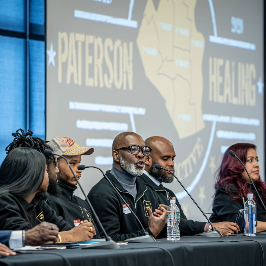 Five panelists seated at an event while one speaker addresses the audience