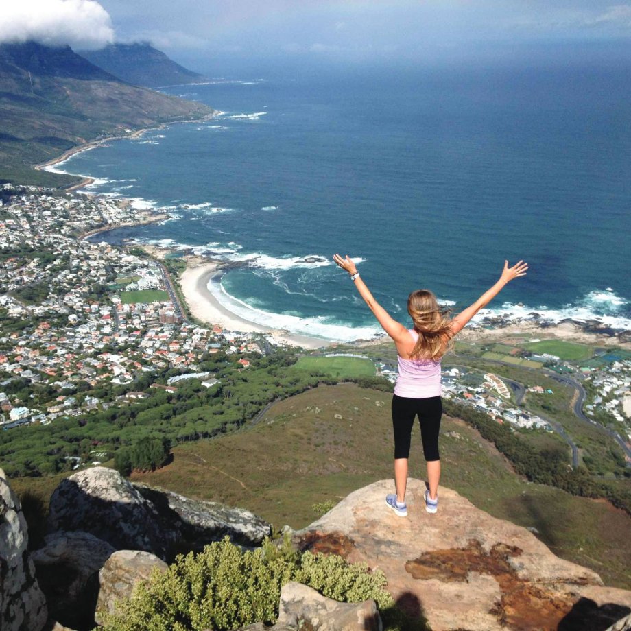 A woman stands on the edge of a tall cliff overlooking a beach