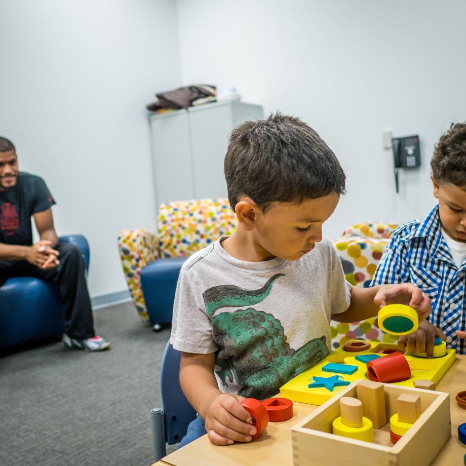 Two children play in an office as their parent watches from afar.