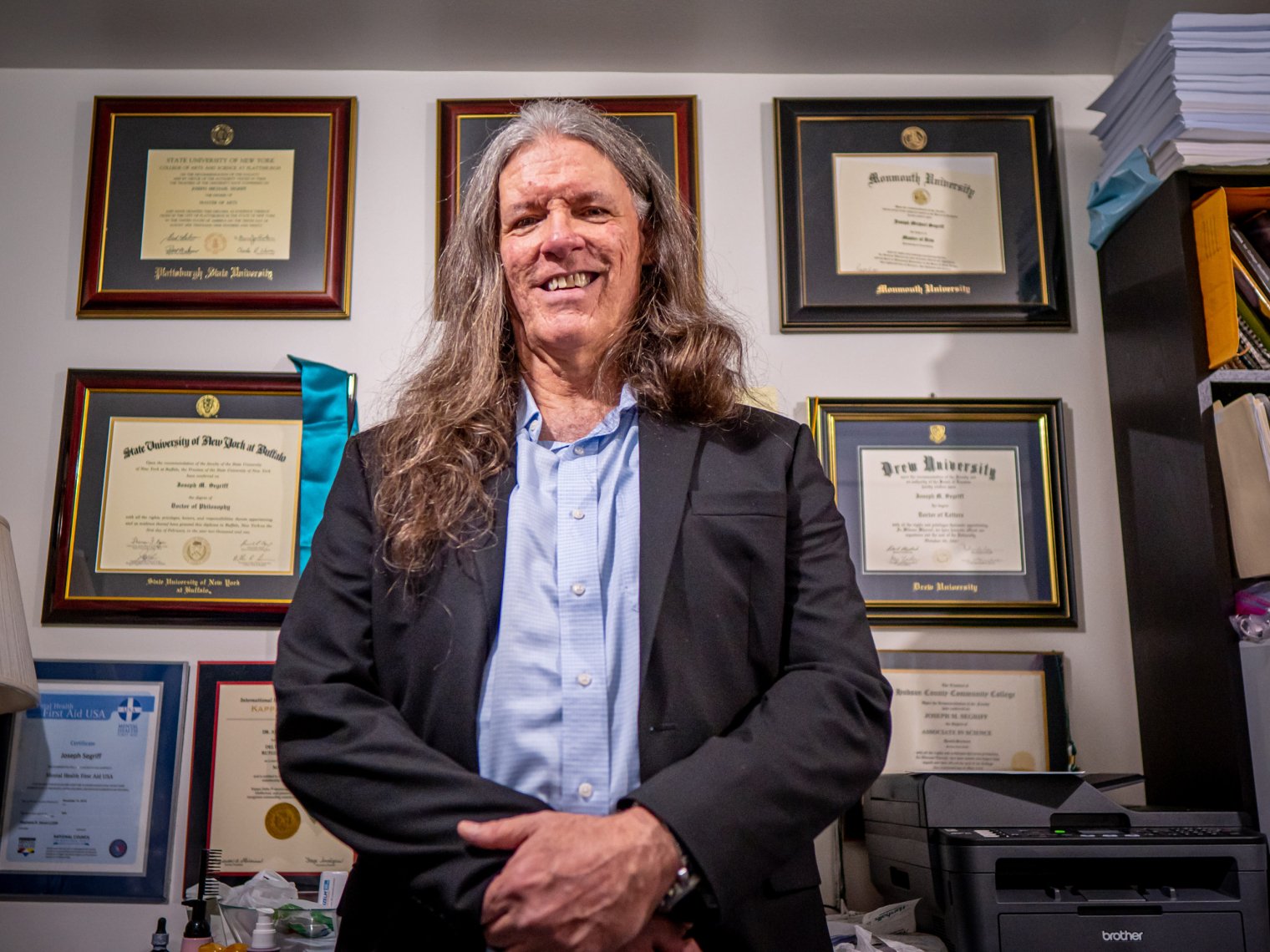A man stands in front of a wall covered with diplomas.