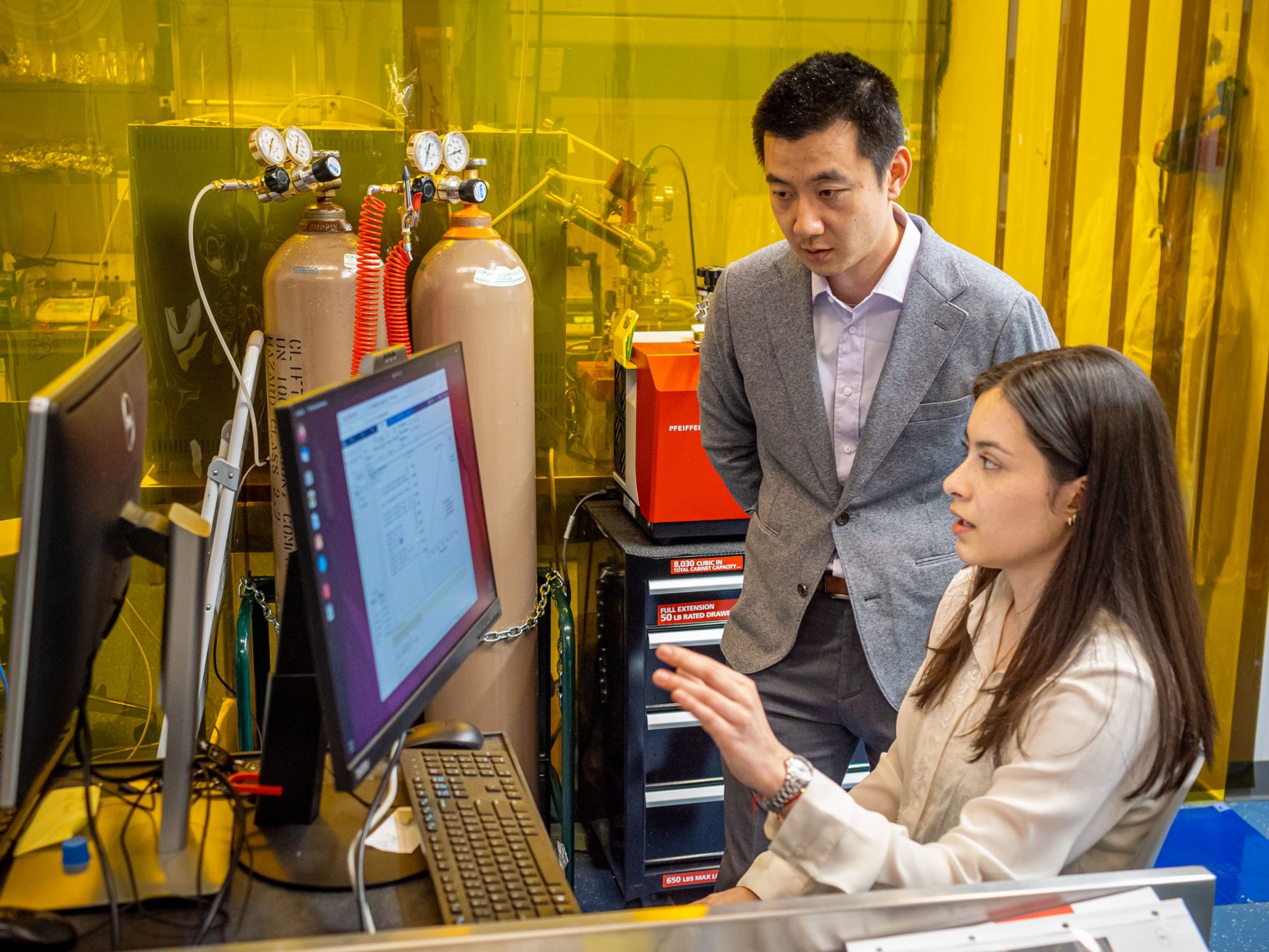 A seated woman gestures at a computer monitor as a standing man looks on.