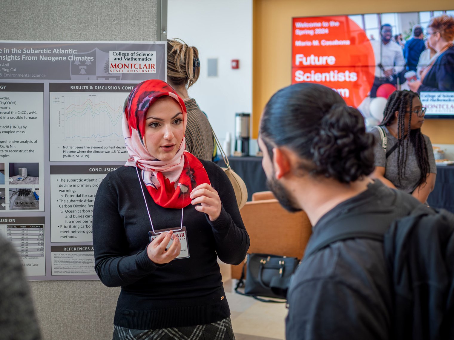 Busra Anil, a Montclair State University student wearing a red and white patterned hijab and black sweater, speaks with a guest during the Spring 2024 Mario M. Casabona Future Scientists Competition. She stands in front of her research poster on carbon sequestration, explaining her findings.