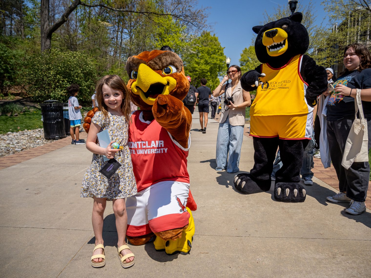 Child poses with hawk mascot from Montclair State University, bear mascot is in the background with a crowd of people.