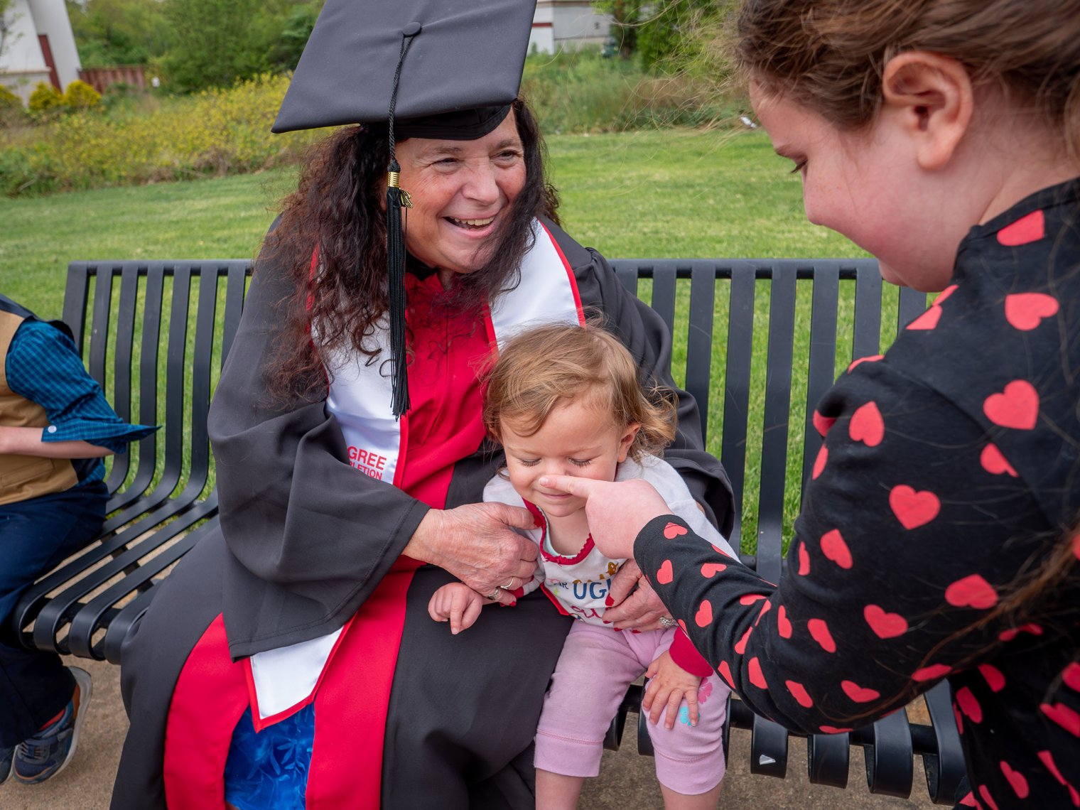 A woman in a cap and gown holds a baby while another woman touches the baby's nose.