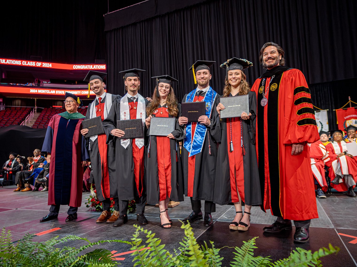 The Povolo quintuplets, wearing graduation caps and gowns, stand together on stage at Montclair State University's 2024 Commencement ceremony. Each holds their diploma while smiling alongside university officials in academic regalia.