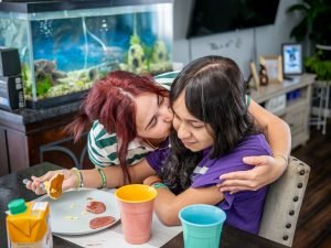 Oscaterin embraces Kamila at the breakfast table, sharing a warm moment to start the day. A plate of eggs, ham and pancakes sits beside Kamila.