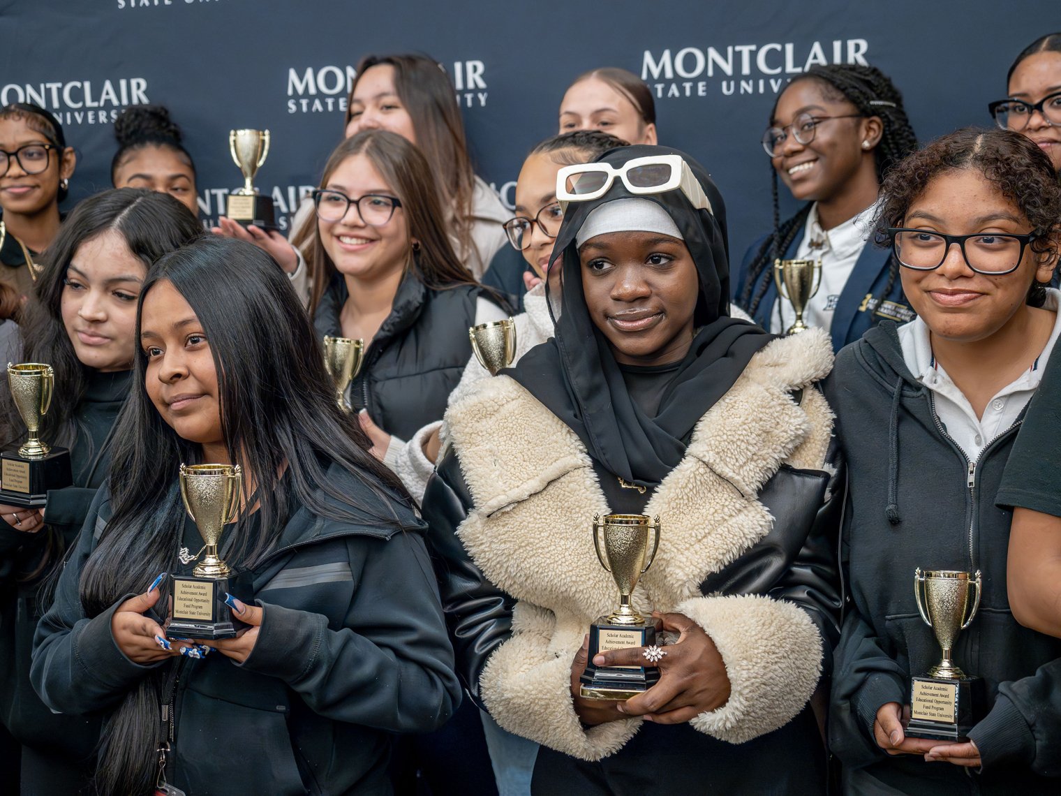 A group of high school students stand together holding leadership awards in front of a backdrop featuring the Montclair State University logo.