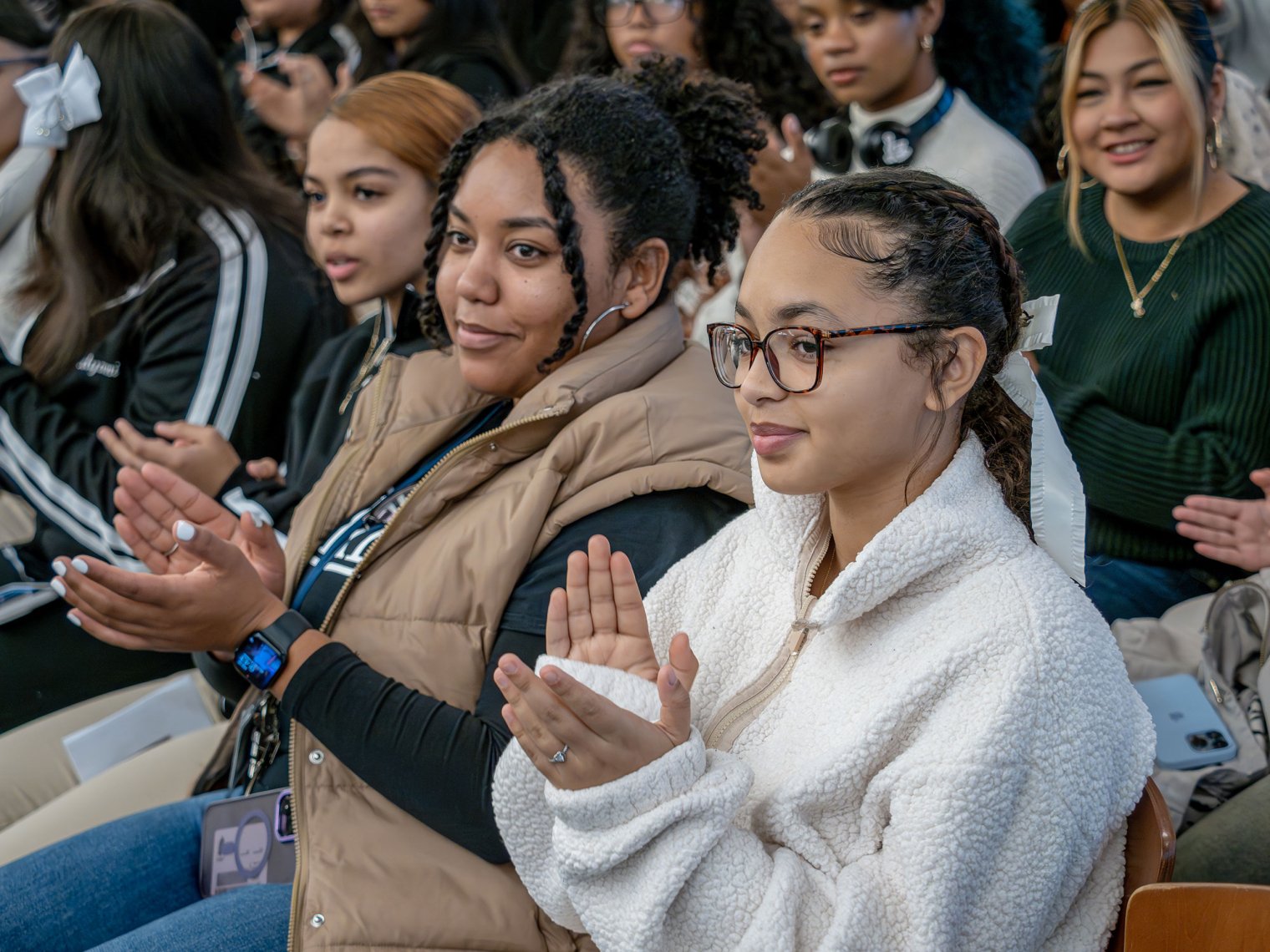A group of high school students sitting in an audience clap their hands during the Women’s Leadership Conference at Montclair State University. The foreground shows two women, one wearing a beige vest and another wearing glasses and a white fleece, both engaged and smiling. The background reveals more attendees focused on the event.
