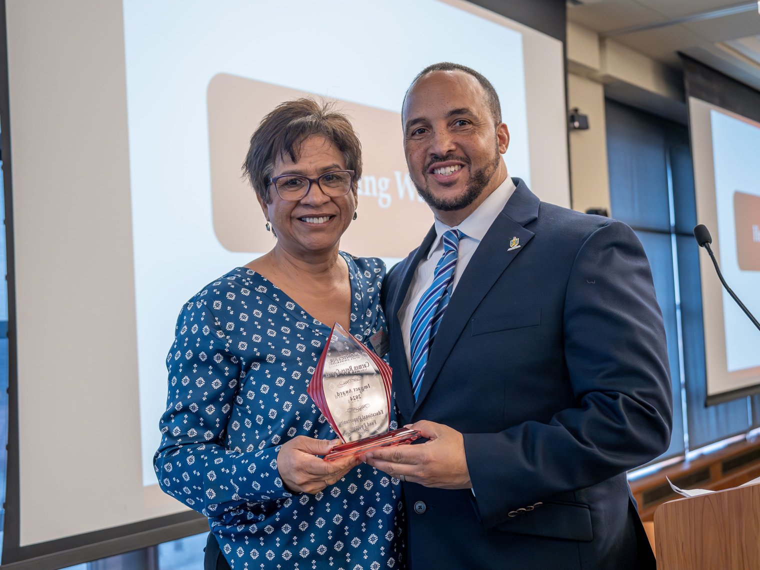Carmen Reyes Cuevas, wearing a blue patterned blouse, holds an award as she poses with EOF alumnus Rey Acevedo, who is dressed in a suit and tie. Both are smiling in front of a presentation screen at the event.