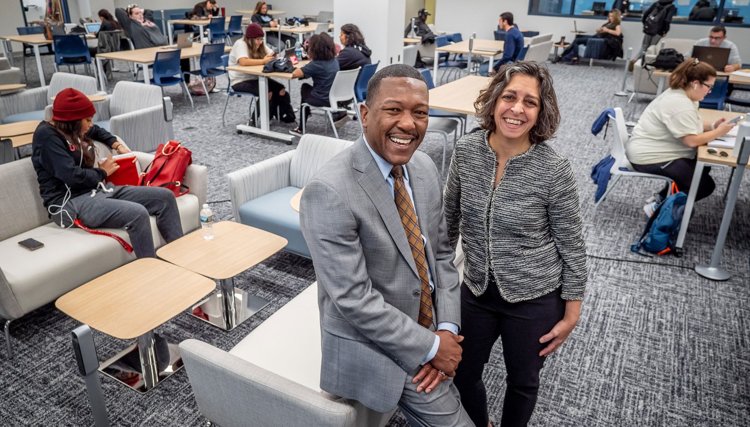 David Hood and Danielle Insalaco-Egan posing on lounge furniture in front of a room filled with students