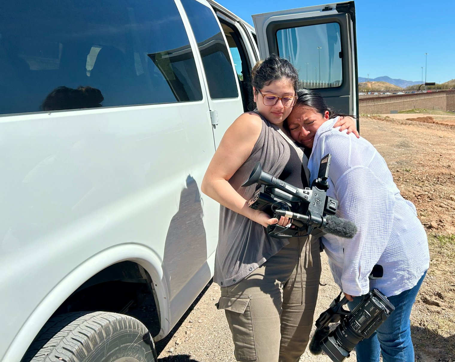 A student holding a video camera hugs another student in the desert.