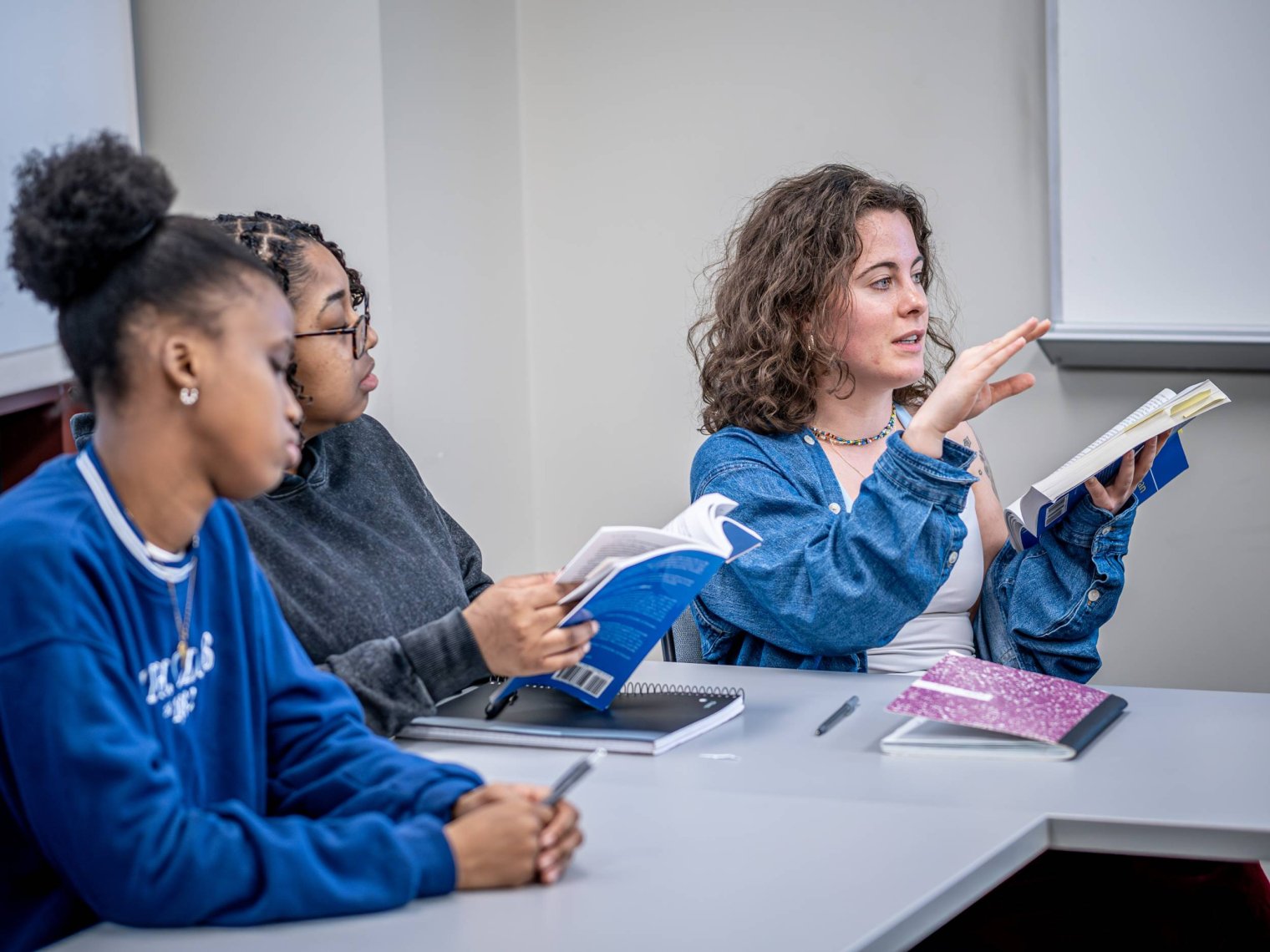 Three students sit in a classroom, two hold books, one is speaking