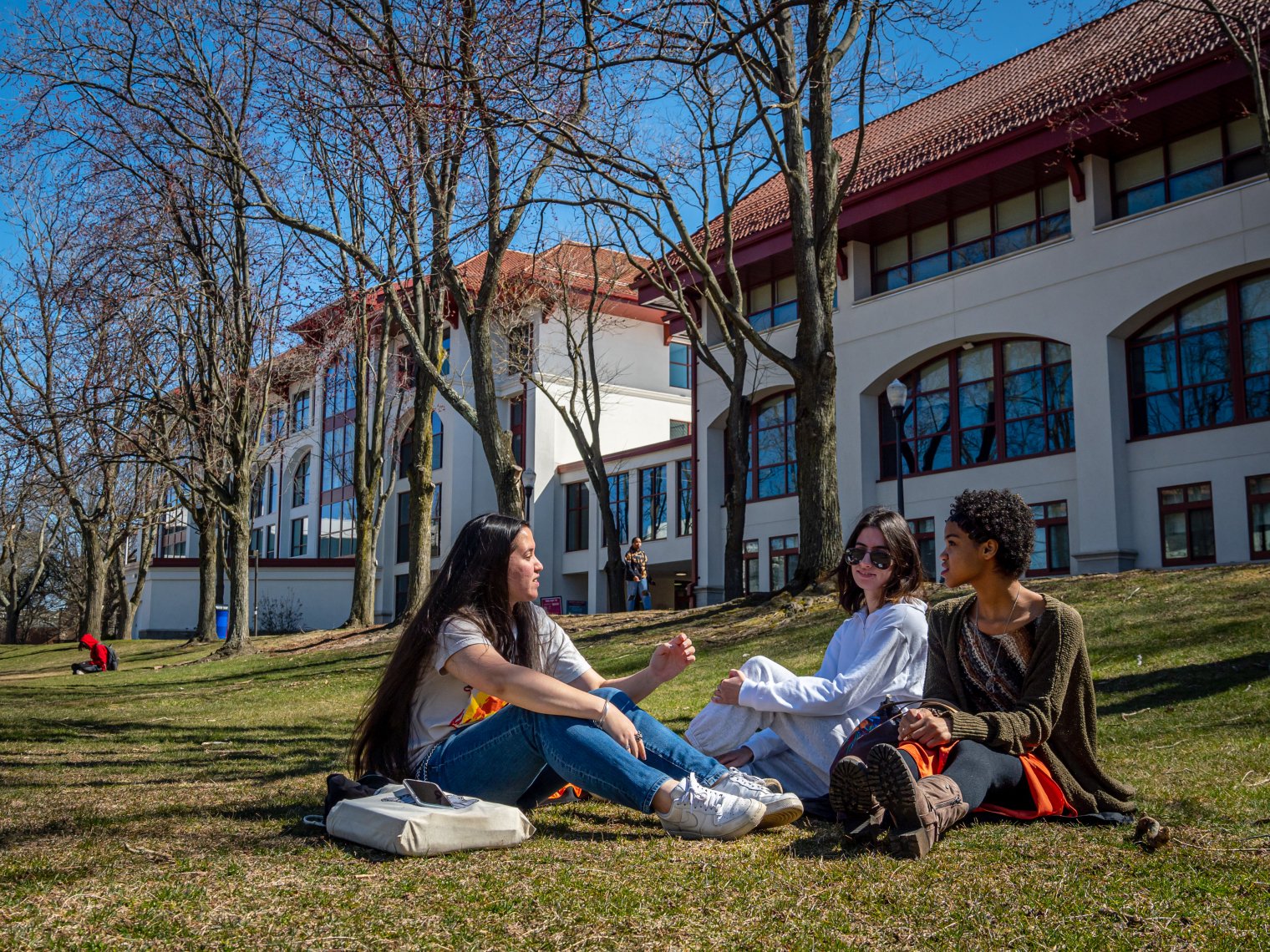 A group of students sitting outside on the grass on Montclair's campus. A campus building is in the background.