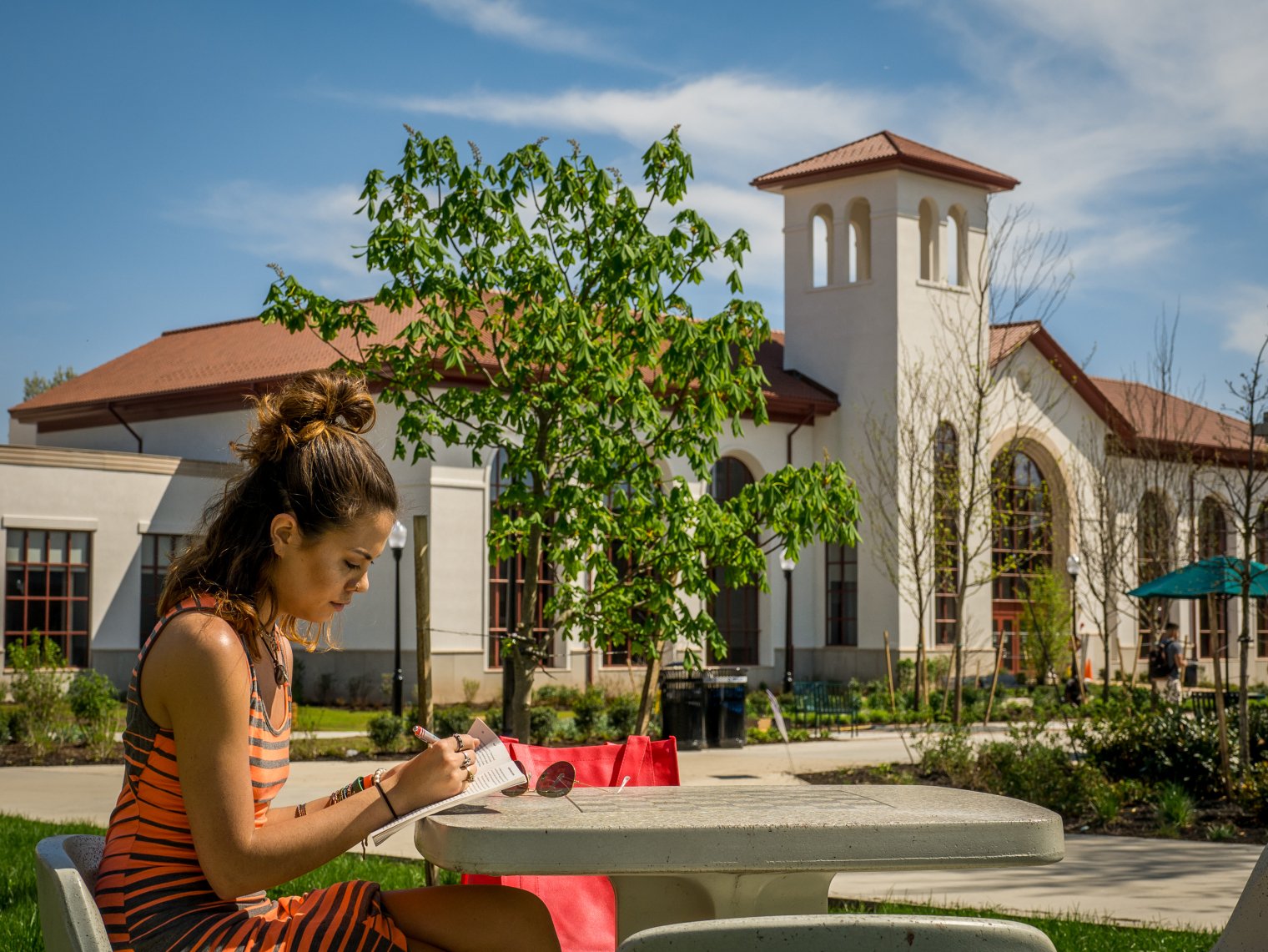 A student sitting at a table outside and writing something in a notebook. Montclair's School of Communication and Media in the background.