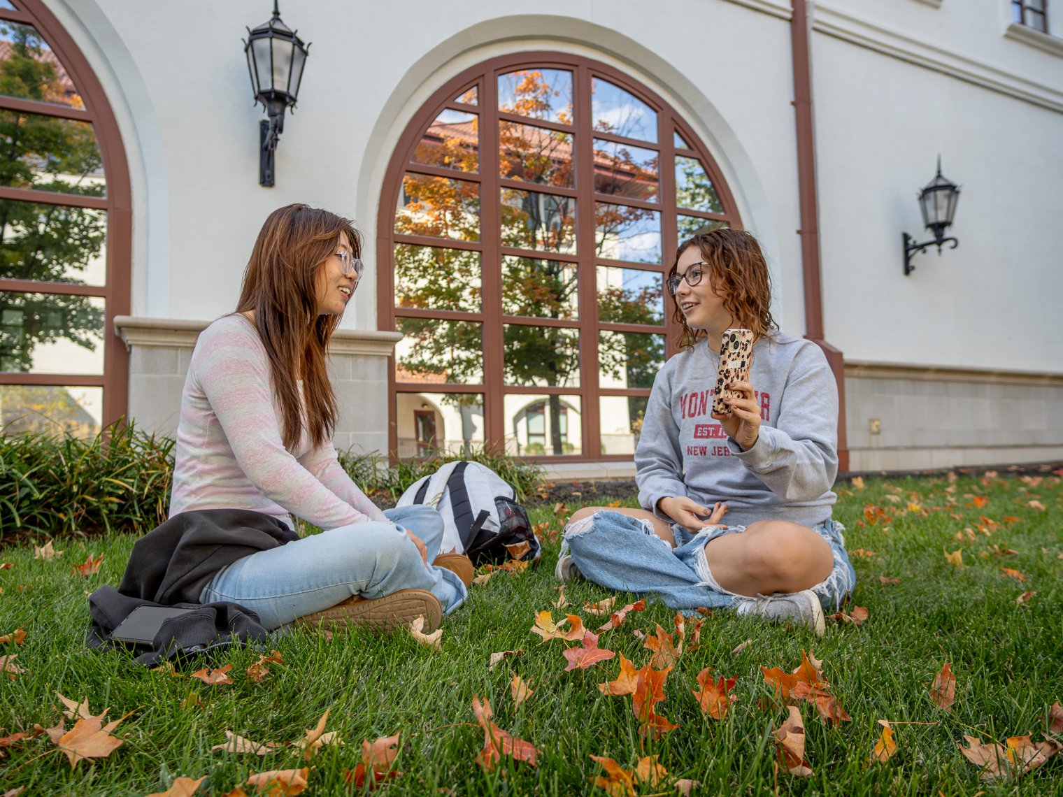 Two students sitting on the grass in front of a building. Fall leaves on the ground.