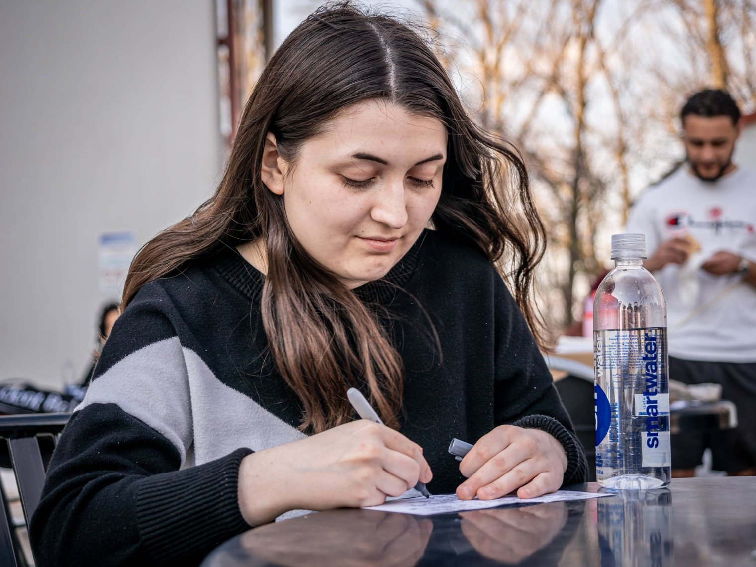 A college student sits at an outdoor table and writes on a piece of paper