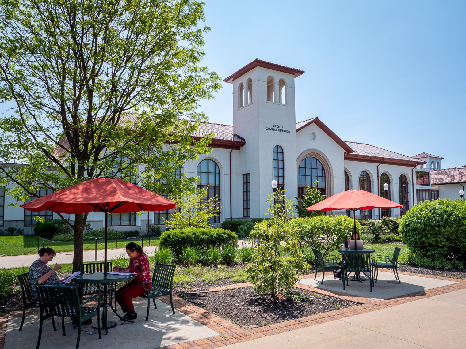 People work at tables with red umbrellas outside of the library on a summer day.