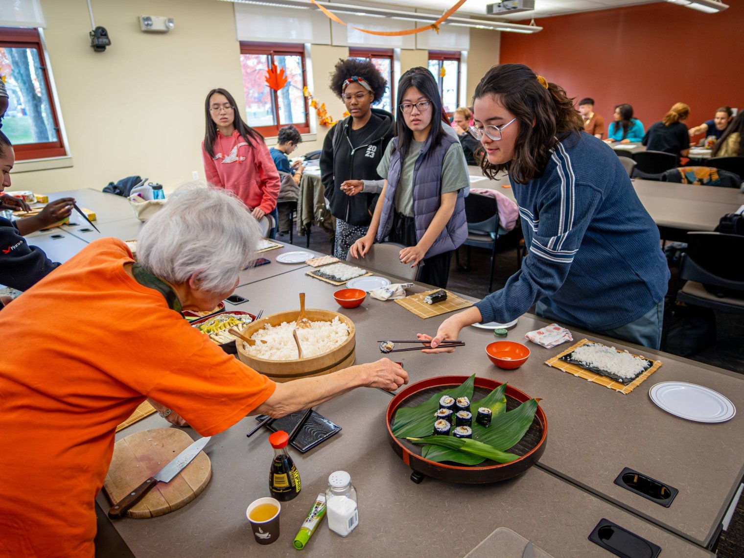 A group of college students watch a woman demonstrate how to make sushi