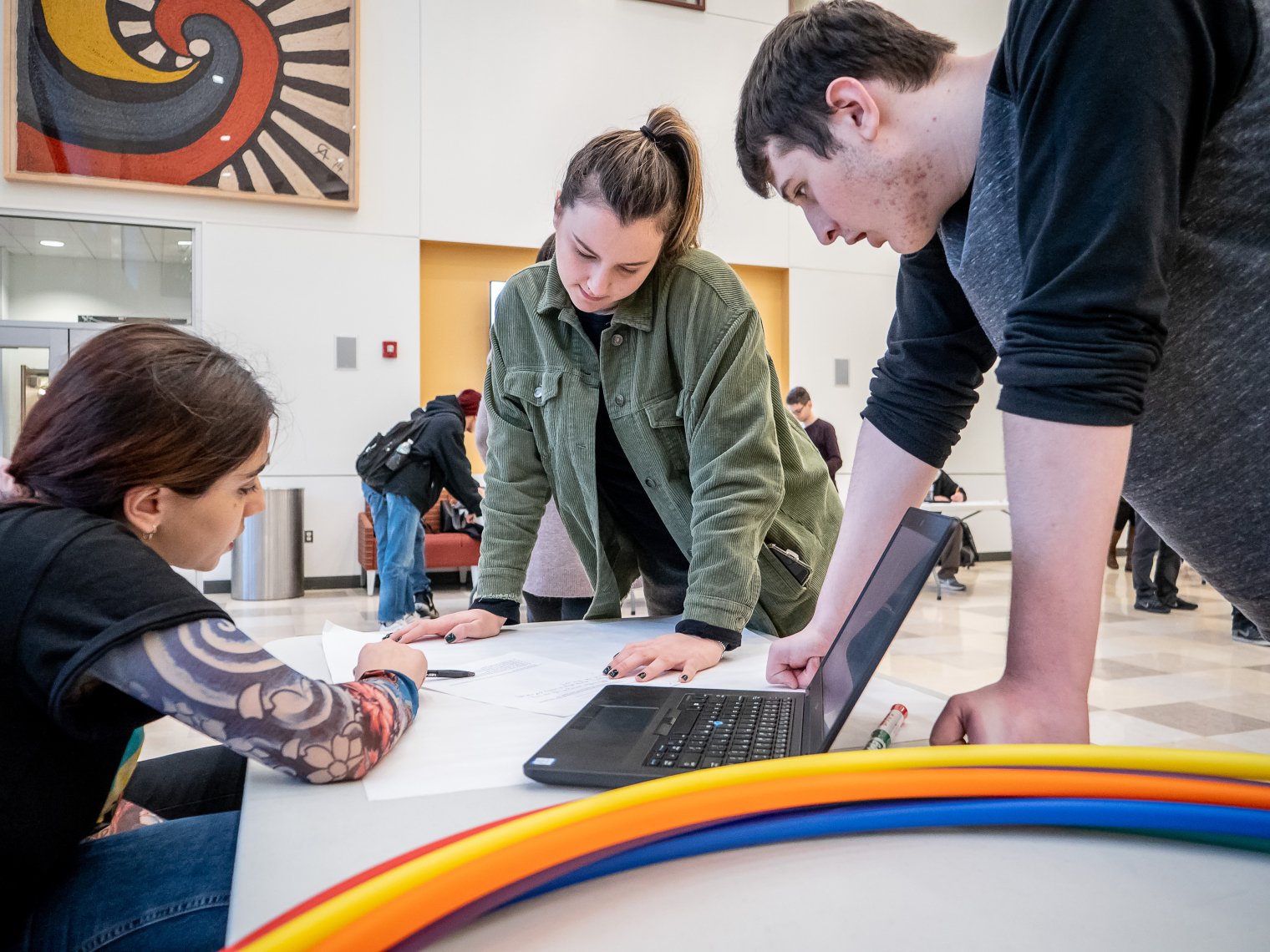 Three students discussing around a table with a laptop and hulahoops joops