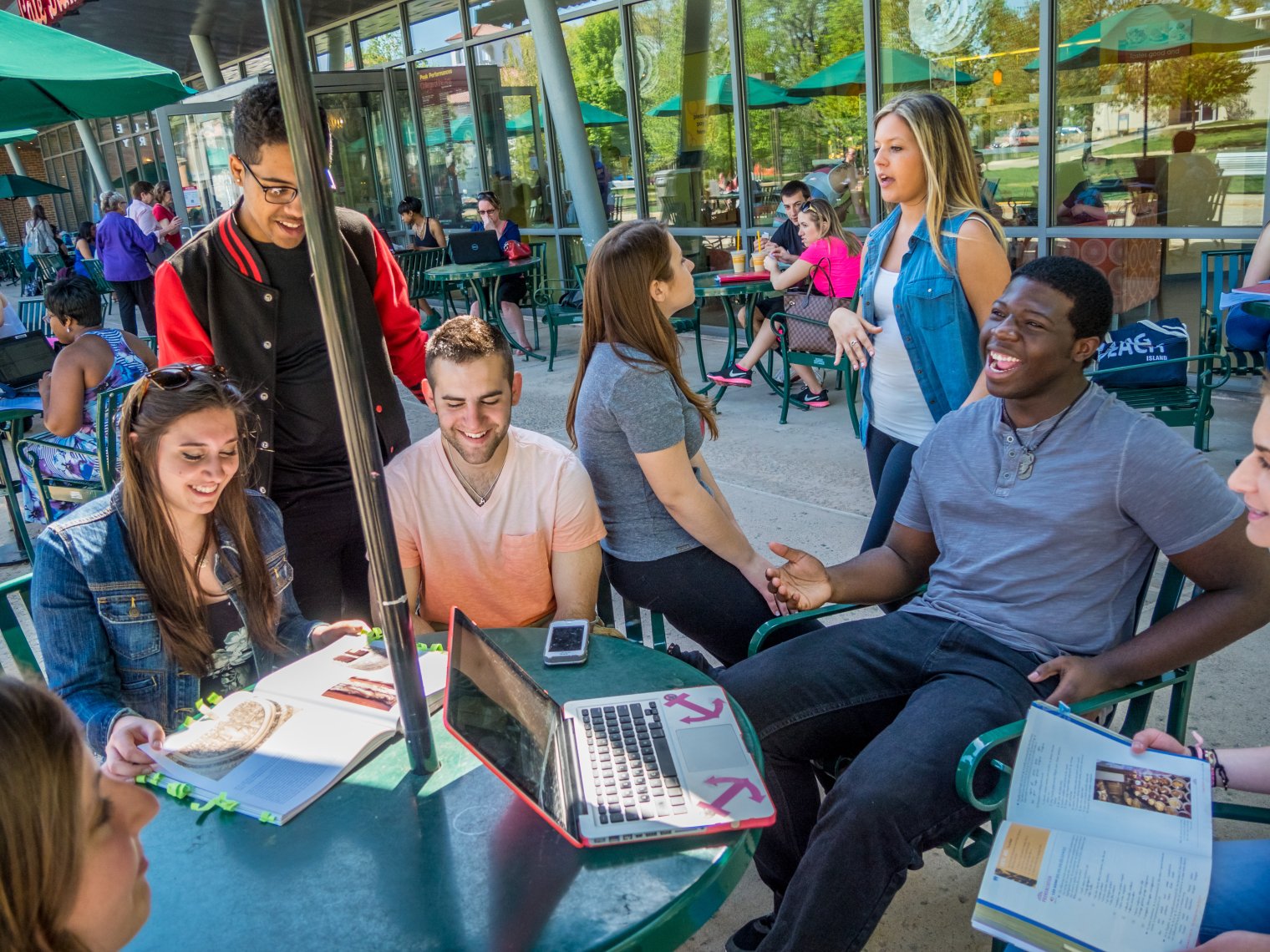 A group of college students sit around an outdoor table under an umbrella with laptops and books out