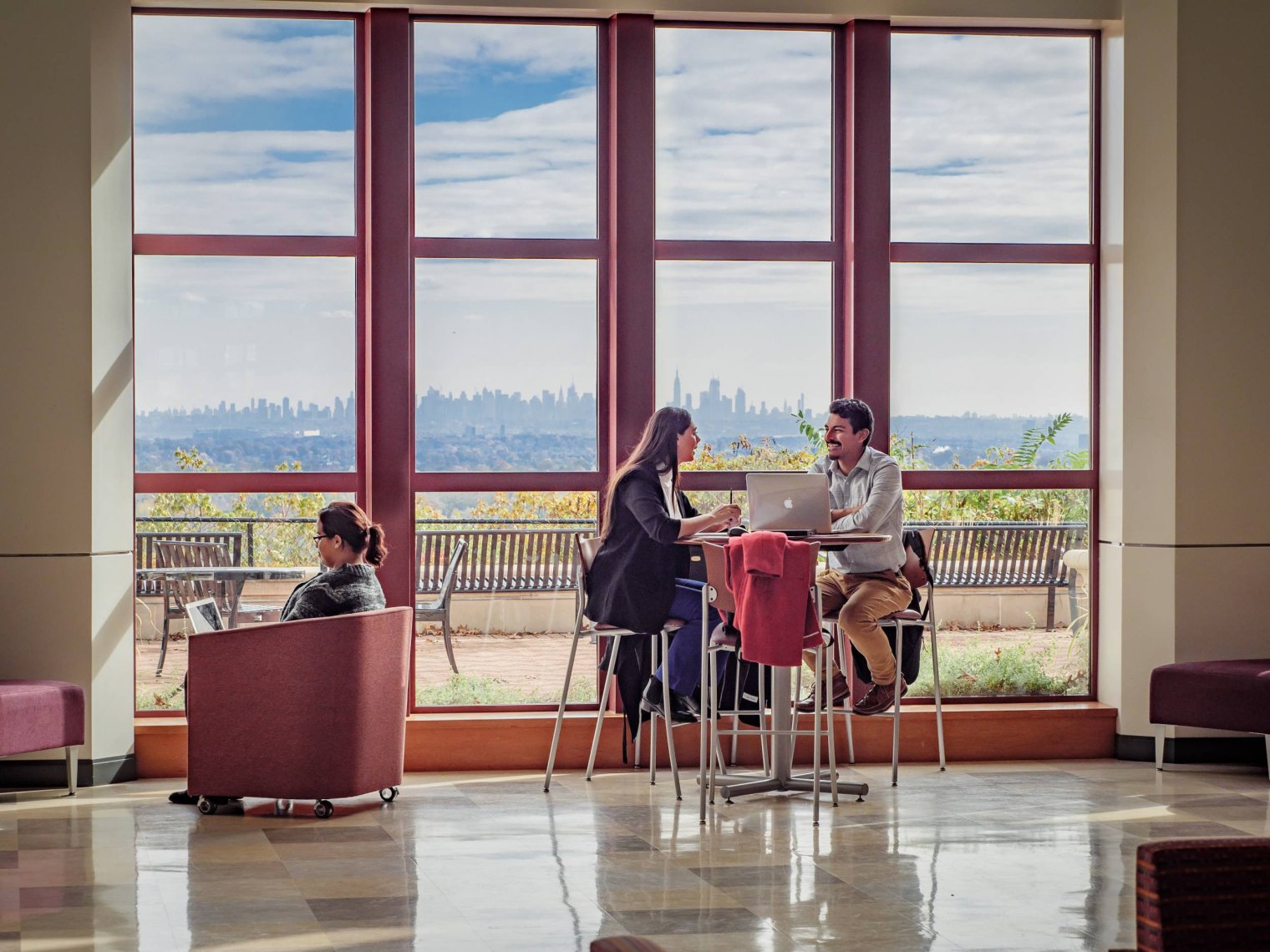 Three students sit inside by a window overlooking the New York City skyline
