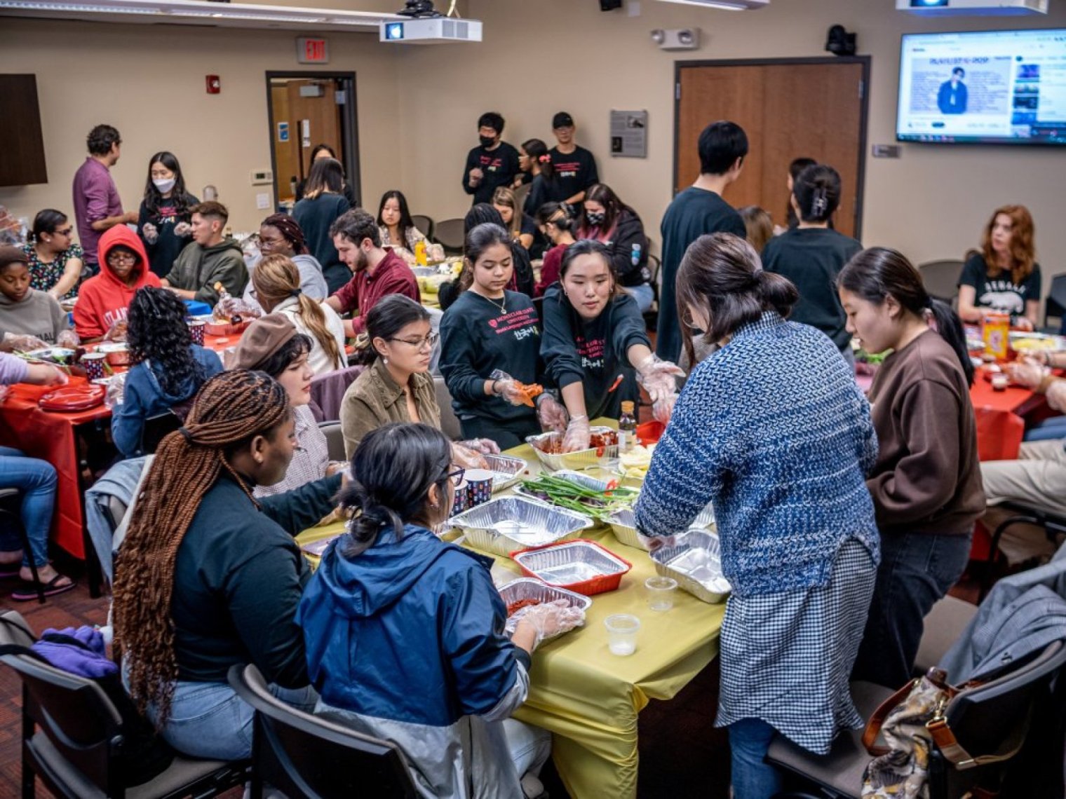 A woman fills serving trays with kimchi as a gorup of college students sit around the table and watch