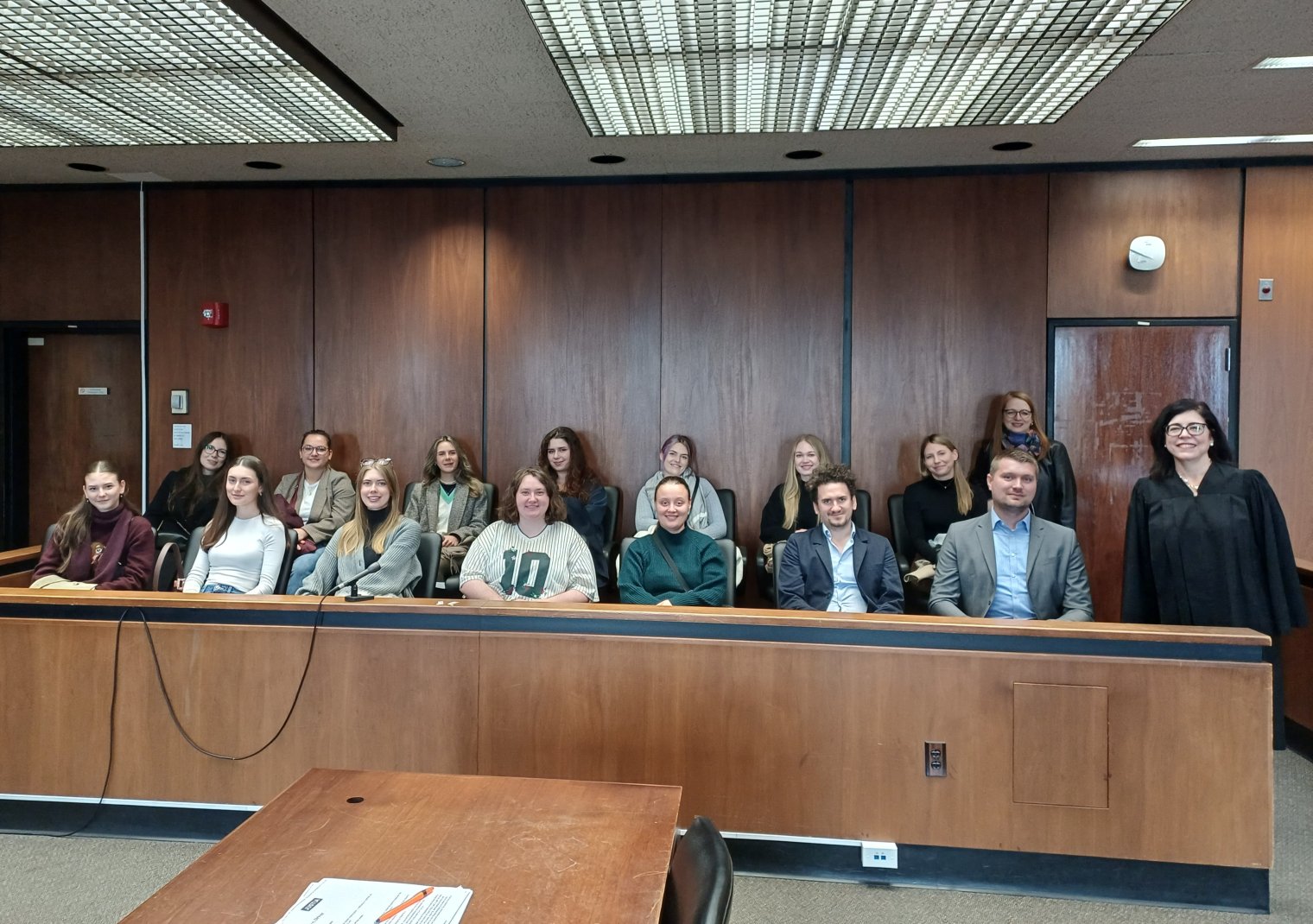 A group of college students sit in a jury box and pose with a judge in black robes