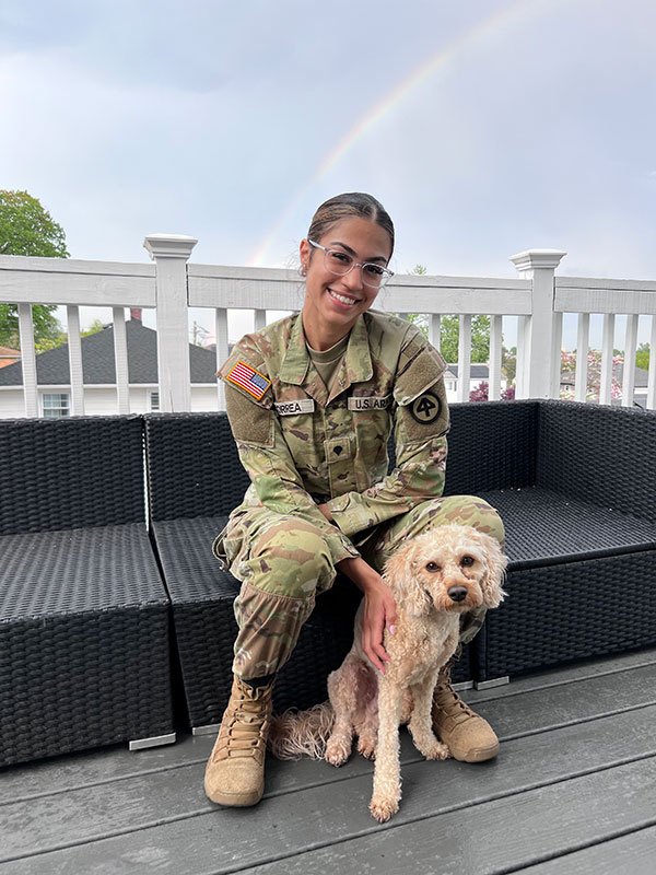 A woman in a National Guard uniform sits outside with a dog, a rainbow is in the background