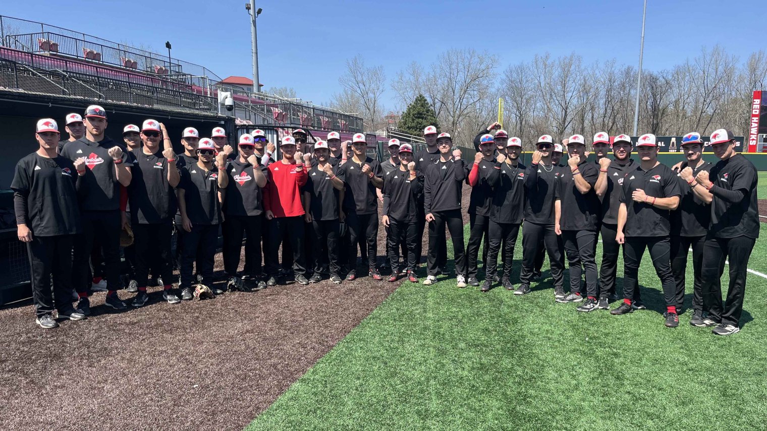 A college baseball team, all dressed in black uniforms, stand in a half-circle on a baseball field