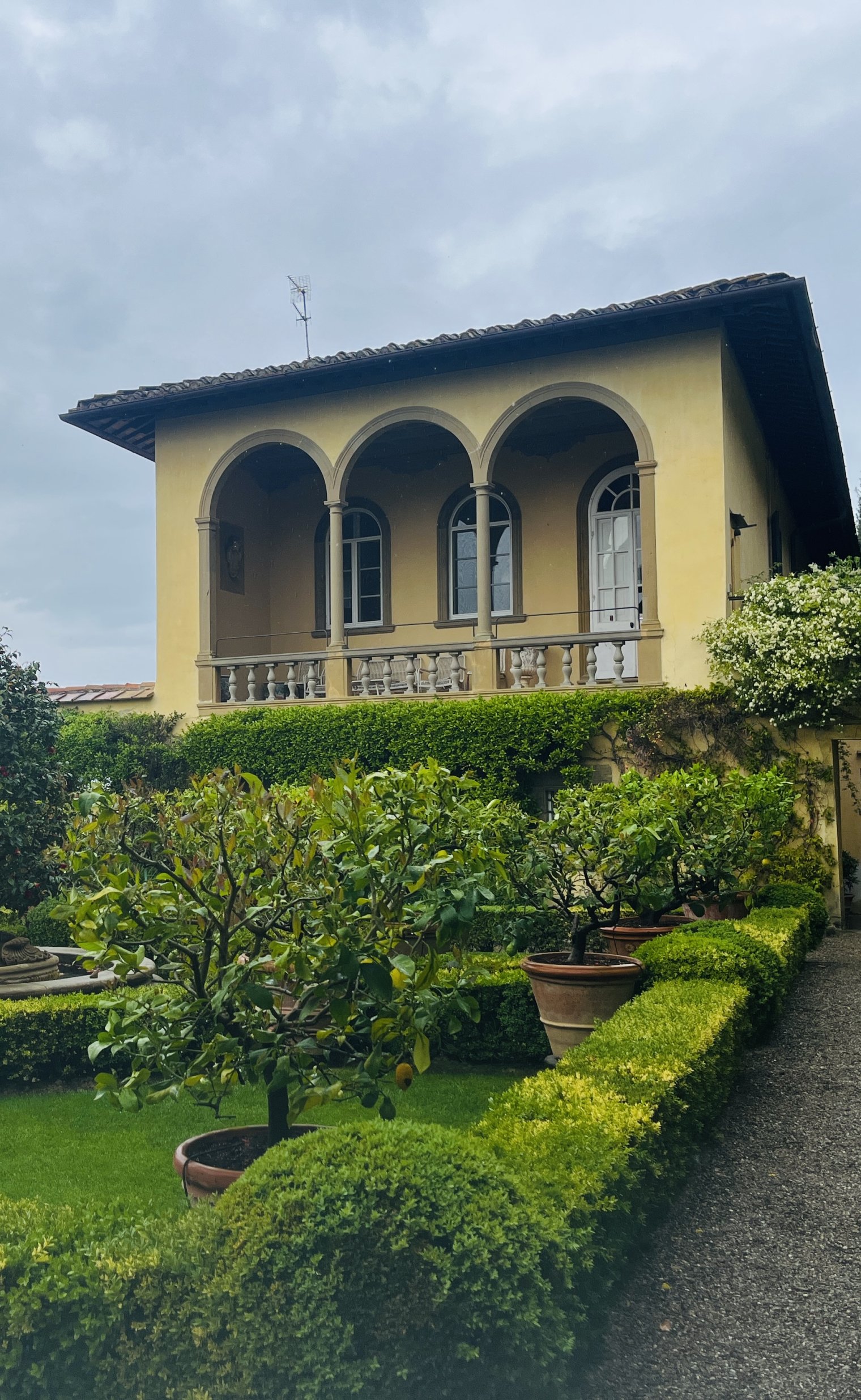 Yellow building in Italy with a patio, potted plants, and manicured hedges on an overcast day