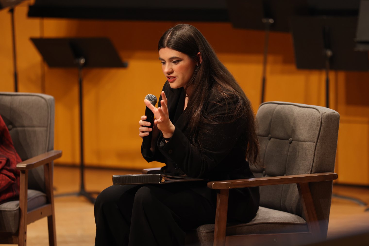 A choir singer sits in a chair on stage, holding a microphone and addresses the audience