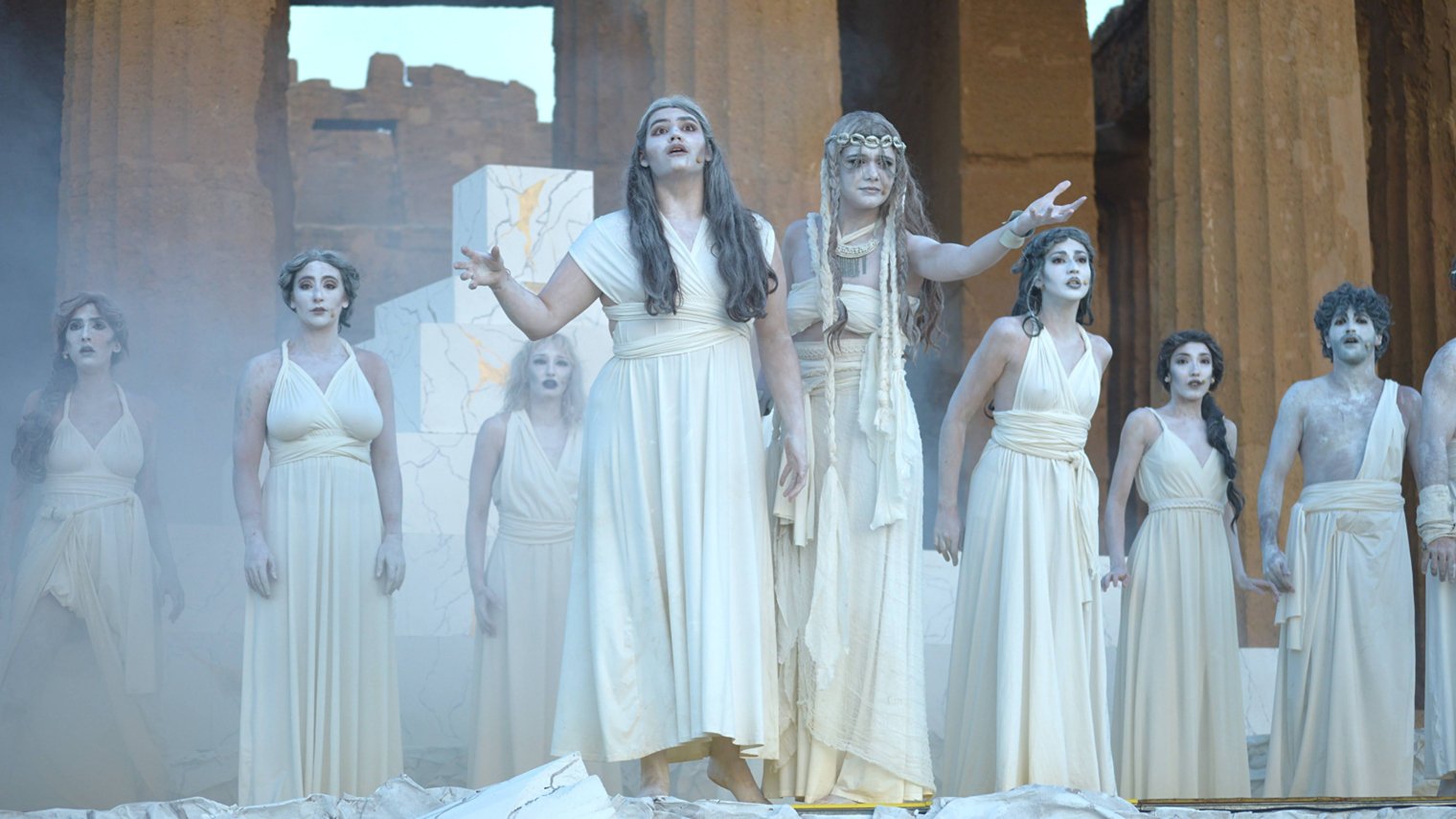 Three students dressed in white toga-style costumes performing the musical Agrigento at an ancient ruin in Italy with large stone columns in the background