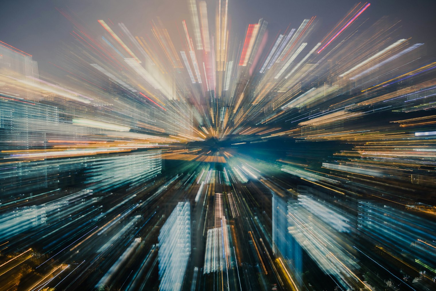 An abstract, long-exposure photo of a city night-time skyline with a radial zoom blur. Bright streaks of white, yellow, and blue light burst outward from a dark center.