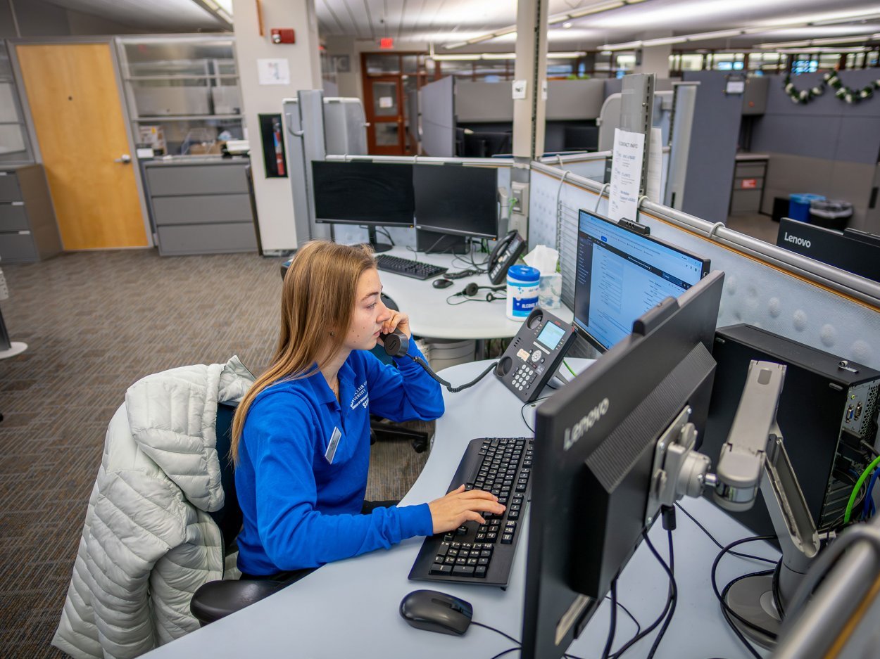 A student working on campus. She is sitting at a computer with her hands on the keyboard and she is on the phone.