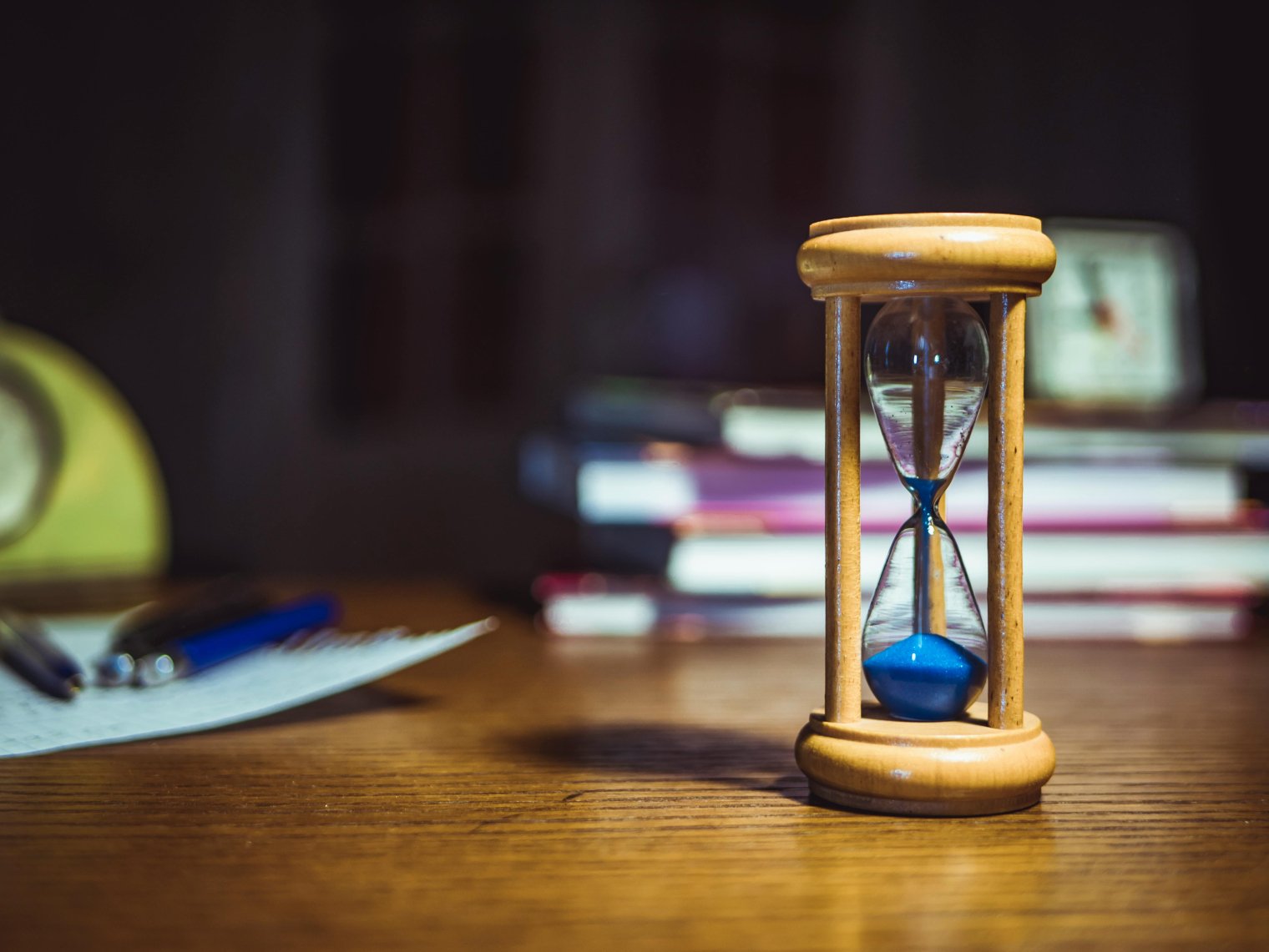 An hour glass on a table in front of a stack of books with a sheet of paper and pens to the back left