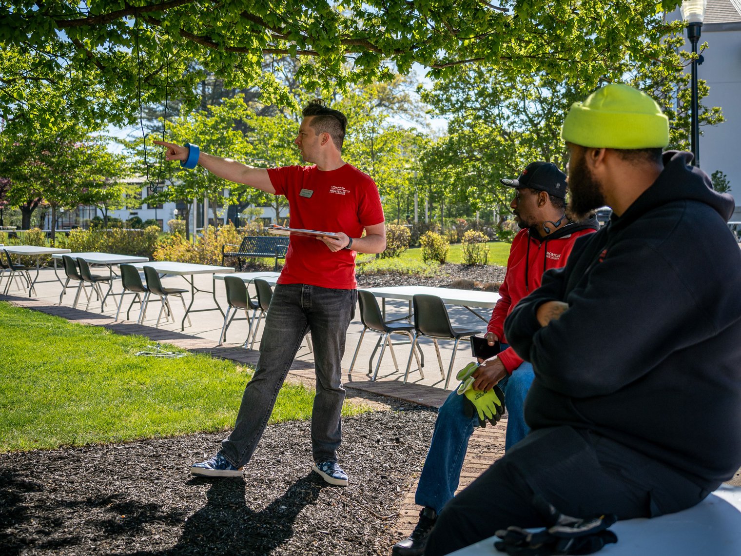 On a sunny morning under leafy green trees, Casey Coleman stands holding a clipboard and points across campus while two Facilities staff members sit on the edge of a table listening.