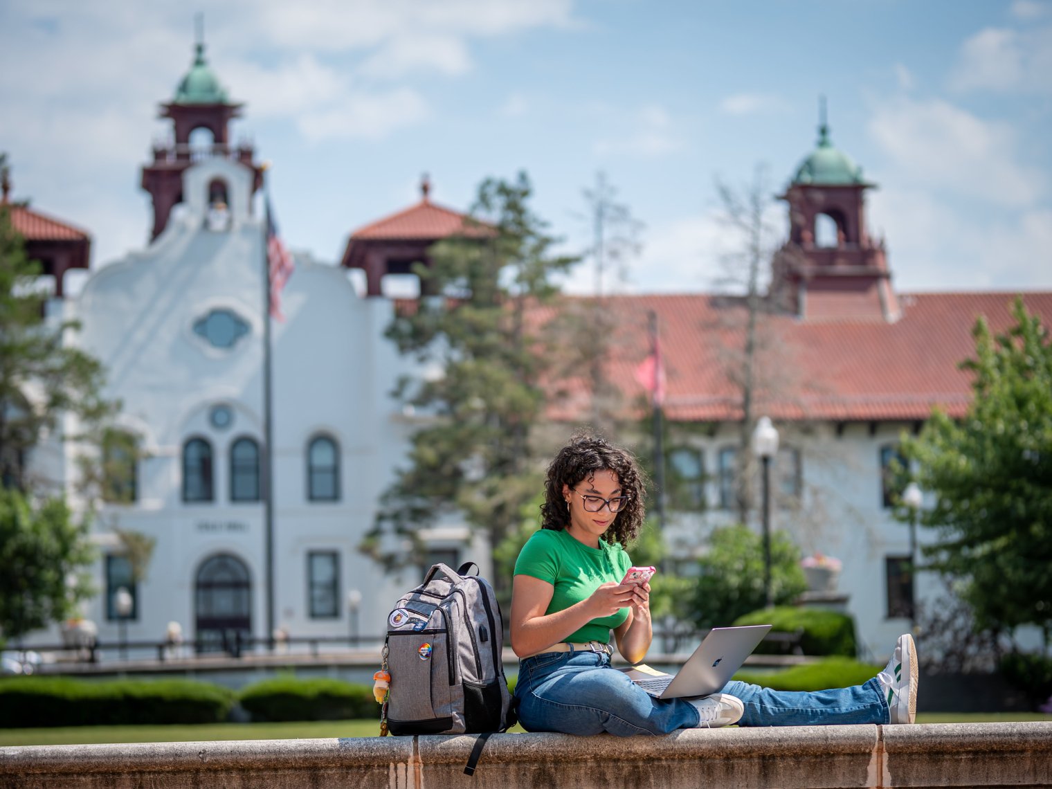 Woman sitting a ledge with laptop, phone, and bookbag. 