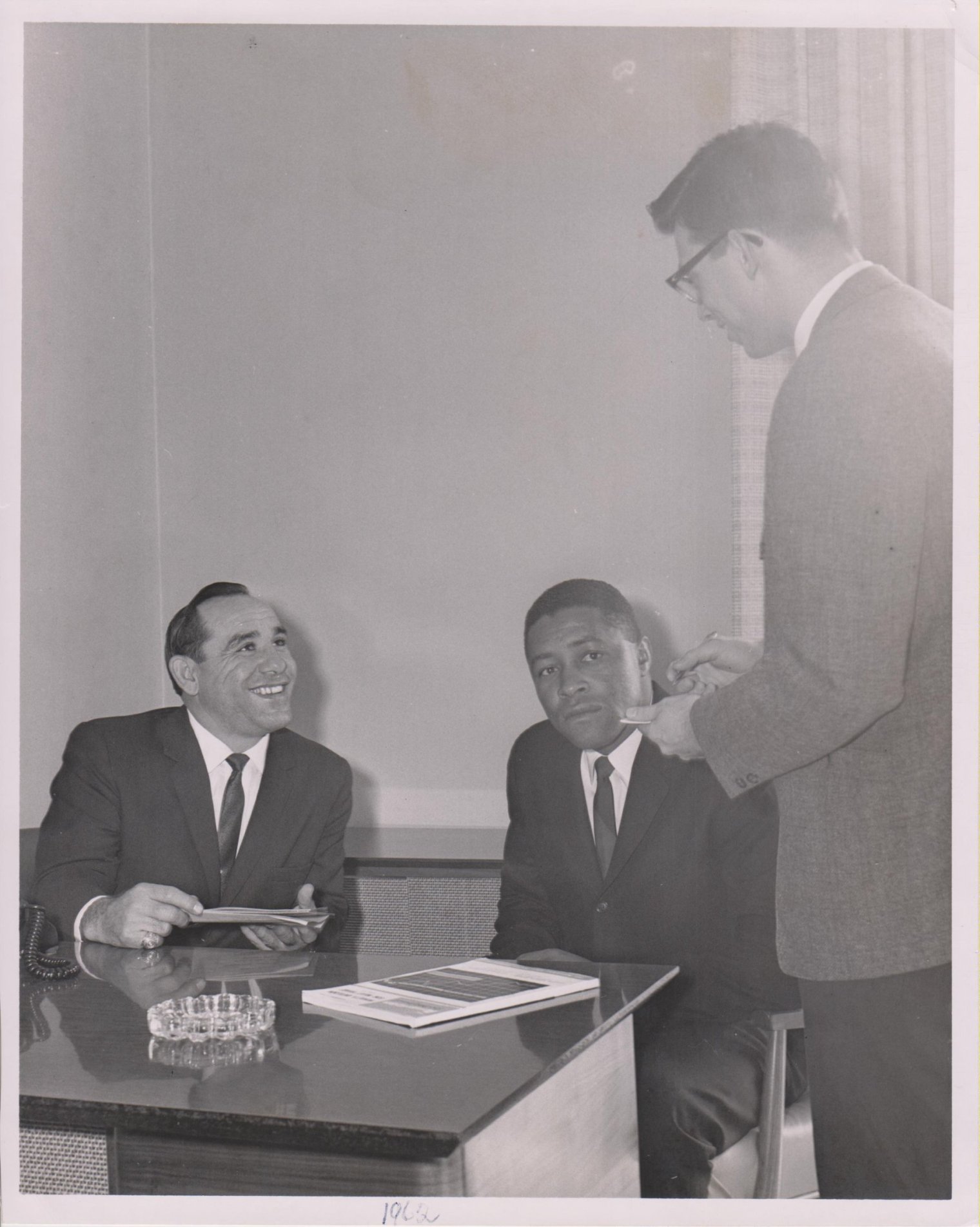 A black and white photograph of a man standing and talking to Yogi Berra and Elston Howard