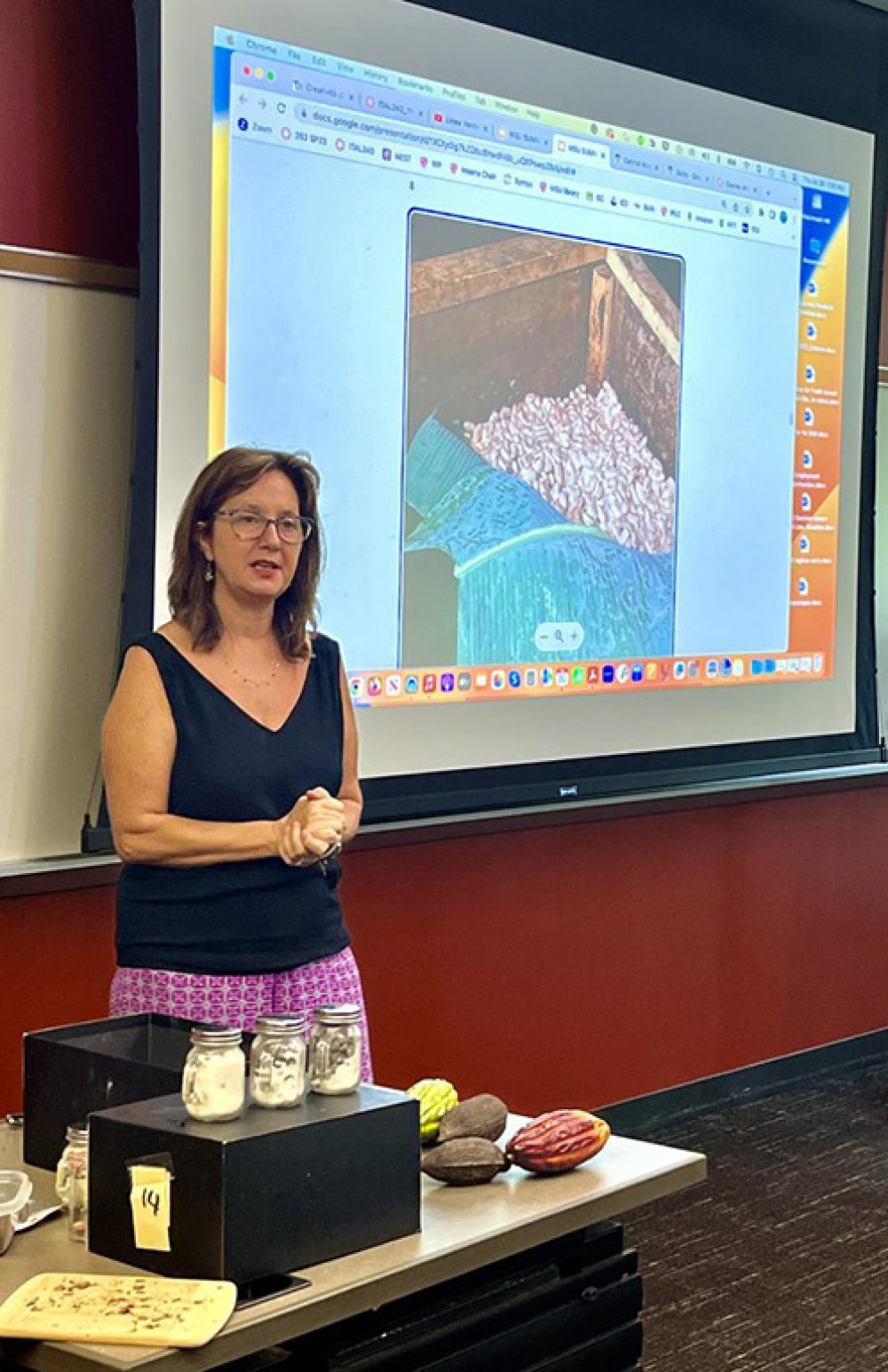 A woman stands in front of a projector screen and delivers a presentation
