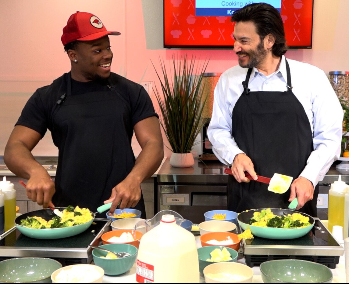 Student and President dressed in black aprons smiling to each other as they stir their food in their pans.