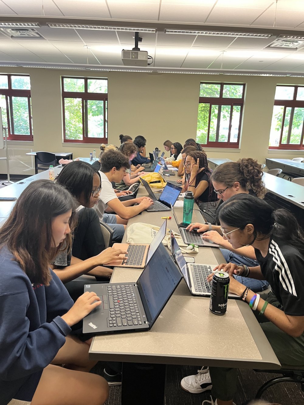 A class of college students sit at a long table and type on their laptops