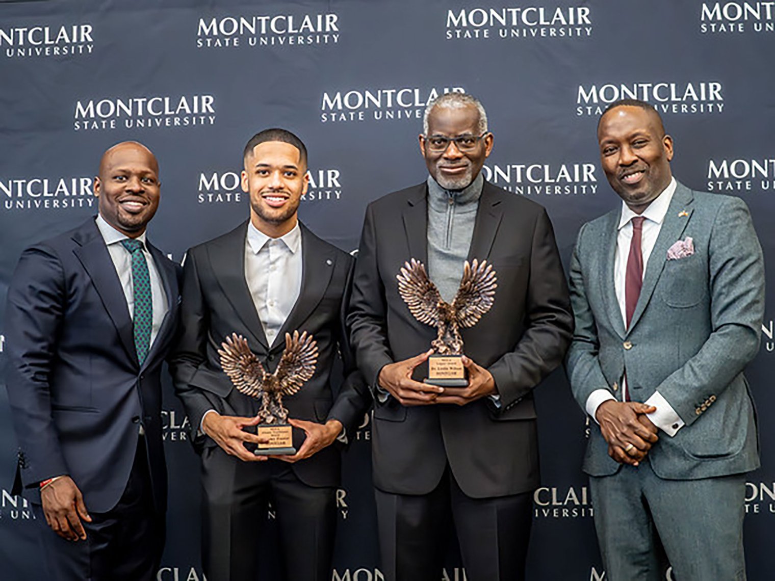 Four man in suits are posing, two in the middle are holding awards.