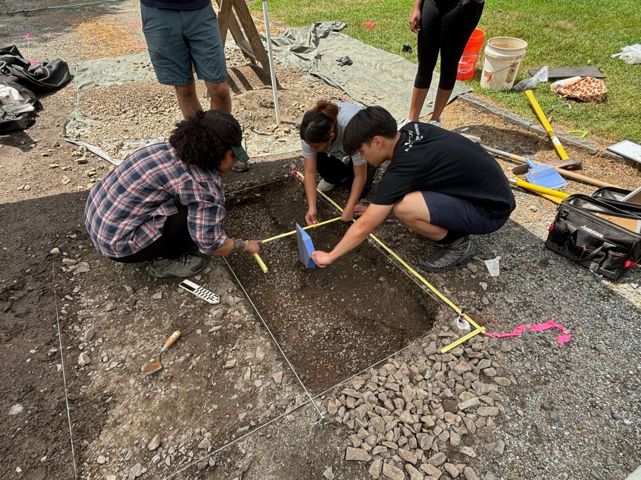 Three college students kneel over a hole in the soil holding tools and a tape measure
