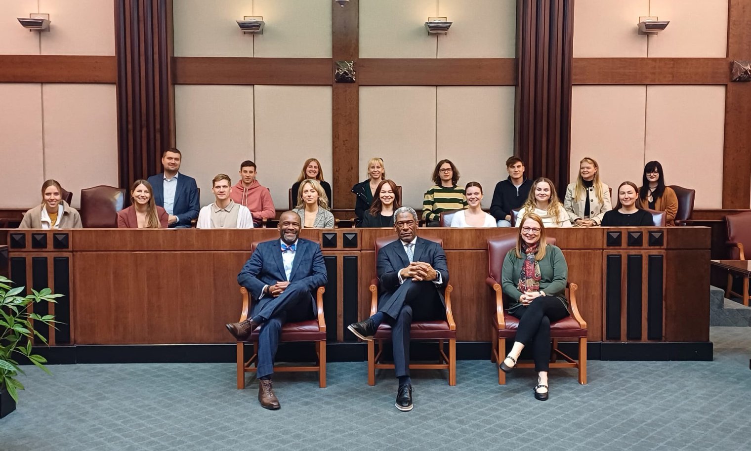 Three people sit in chairs and pose in front of a jury box in a court room