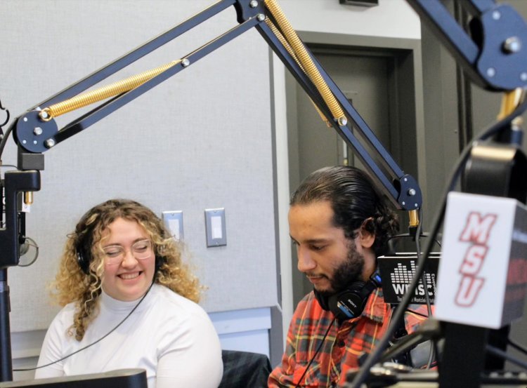 Two people sit at a microphone in a radio studio