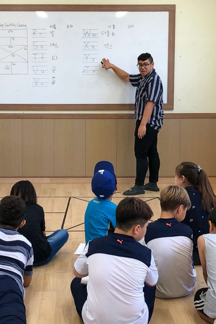 A college student stands at the front of a classroom and points to the lesson on the board